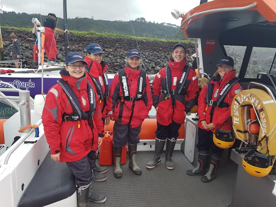 Five young people standing in boat wearing red jackets, caps, with life jackets on 