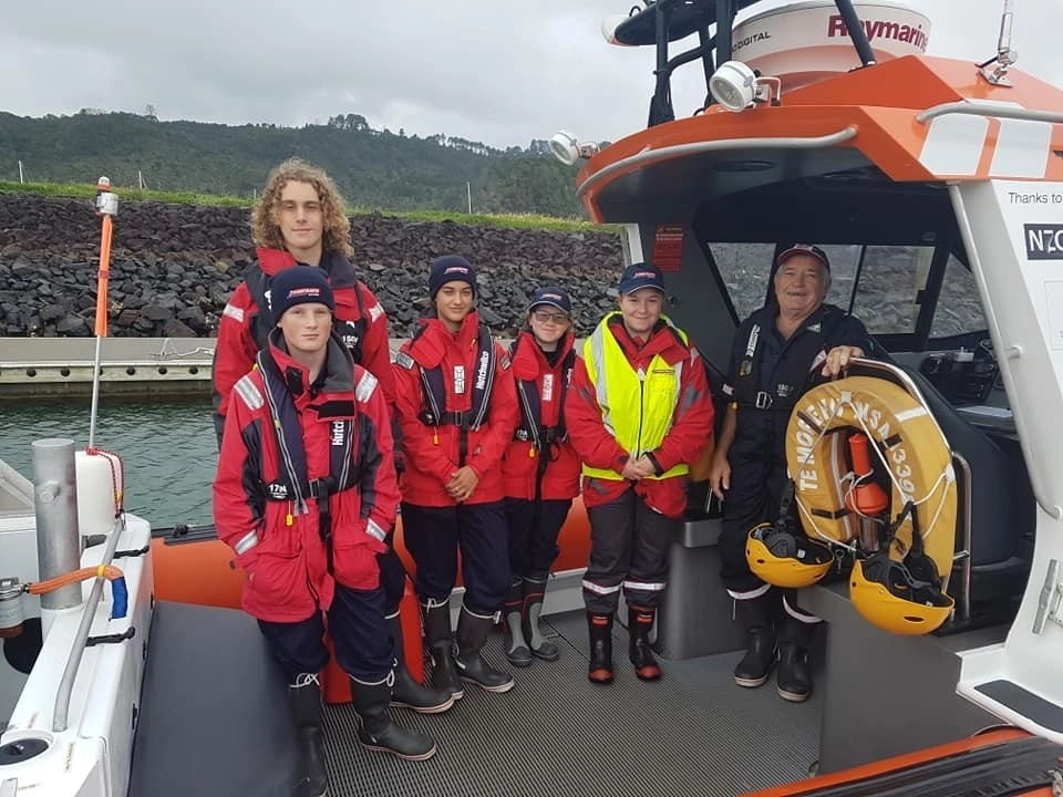 Five young people and skipper standing in boat wearing red jackets, caps, with life jackets on