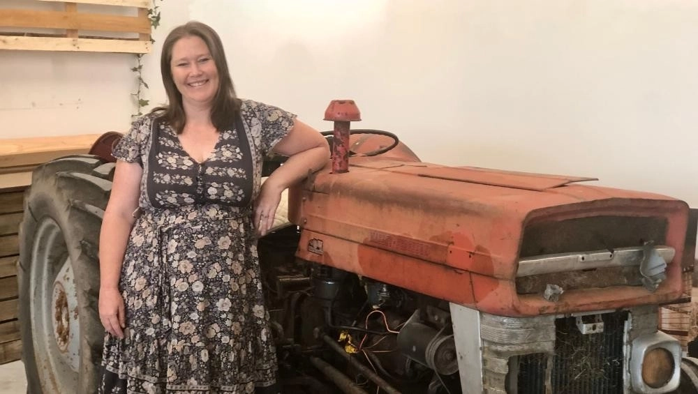 Woman (Lisa Gearon ) standing beside old red tractor.